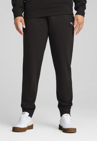 Svarta mjukisbyxor med en slät yta och elastiska muddar, prydda med en liten vit logotyp på vänster höft. Kombinerat med vita sneakers.