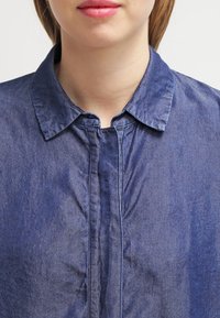 Camicia in denim blu con colletto classico, patta frontale e texture sottile. Presenta un tessuto morbido e una vestibilità comoda.