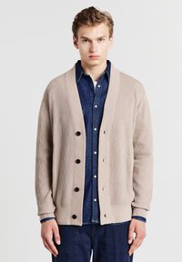 Beige gestreifter Cardigan mit einem Schalkragen und sechs schwarzen Knöpfen. Über einem dunklen Denim-Hemd getragen, das eine lockere Passform zeigt. Weiche Textur.