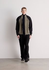 Férfi sportos öltözet, amely fekete és olívazöld cipzáras dzsekit, fekete garbót és laza fekete, sima textúrájú nadrágot tartalmaz.