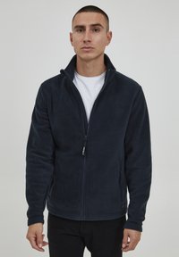 Chaqueta de forro polar azul marino con cuello alto, cremallera completa y bolsillos laterales. Textura suave, diseño ajustado y una sutil etiqueta de la marca en el dobladillo.