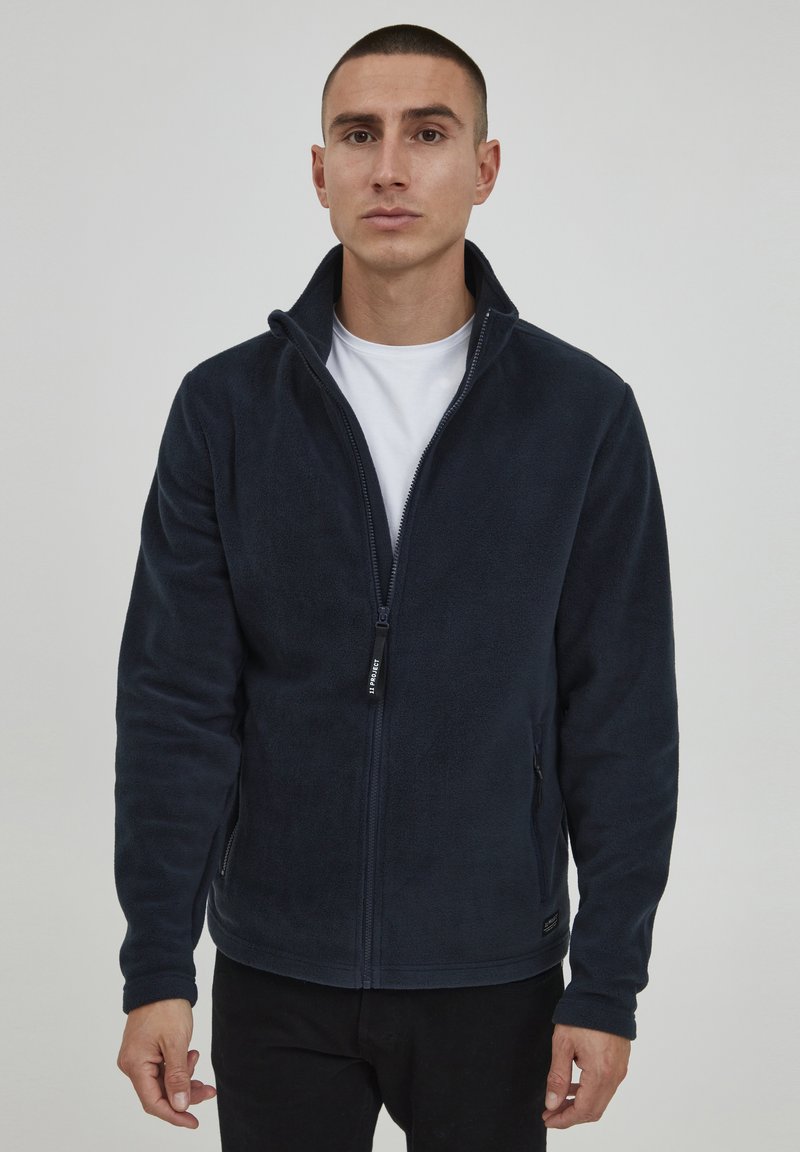 Marineblaue Fleecejacke mit hohem Kragen, durchgehendem Reißverschluss und Seitentaschen. Weiche Textur, taillierte Passform und ein dezenter Markenanhänger am Saum.