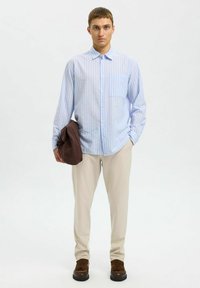 Camisa de botones a rayas azul claro con un bolsillo en el pecho, pantalones beige y zapatos marrones, sosteniendo una chaqueta marrón. Diseño simple y moderno.