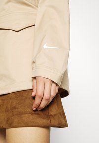 Beige Nike jacket with a white logo, featuring a lightweight fabric and a relaxed fit. Hand rests on a brown suede skirt.