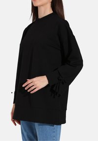 Felpa nera oversize con collo rotondo, maniche lunghe con nastri, e un tessuto liscio e testurizzato. Abbinata a jeans azzurri chiari.