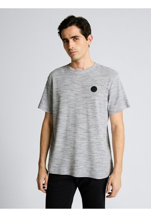 Mann trägt ein hellgraues kurzärmeliges Hemd mit einem schwarzen runden Logo auf der linken Brust und steht vor einem einfarbigen hellen Hintergrund.