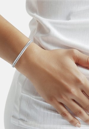 Muñeca con pulsera plateada en espiral, llevada en la mano izquierda, apoyada contra ropa de tela blanca texturizada.