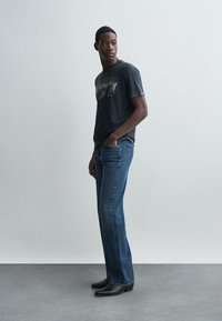 Černé grafické tričko s metalickými akcenty, v kombinaci s modrými džíny rovného střihu. Modelka má na sobě černé boty a stojí na světlém pozadí.