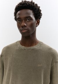 Khaki ribstrik sweater med rund halsudskæring, der har en subtil tekstur og en broderet logo-detalje på brystet.