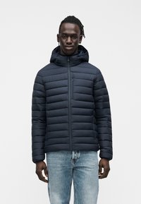 Μπλε σκούρο μπουφάν με κουκούλα, με οριζόντιο σχέδιο καπιτονέ και φερμουάρ μπροστά. Συνδυάζεται με ανοιχτό μπλε τζιν για casual εμφάνιση.