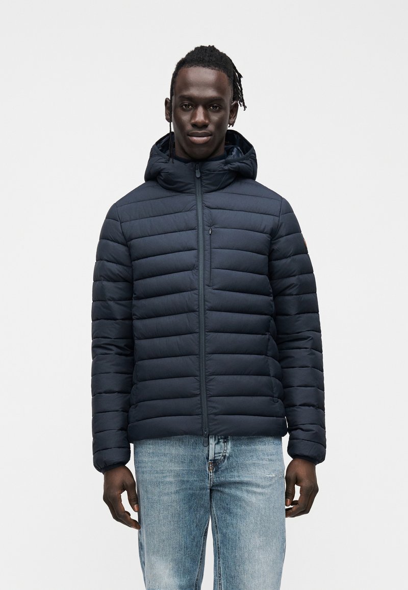 Μπλε σκούρο μπουφάν με κουκούλα, με οριζόντιο σχέδιο καπιτονέ και φερμουάρ μπροστά. Συνδυάζεται με ανοιχτό μπλε τζιν για casual εμφάνιση.