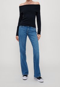 Haut noir à épaules dénudées et manches longues, associé à un jean évasé bleu clair et à des baskets noires aux semelles blanches. Le matériau semble lisse, mélange de coton.