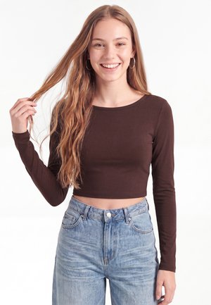Ung kvinde smilende, iført en langærmet mørkebrun crop top og lyseblå højttaljede jeans, holder en tot af sit lange bølgede hår.