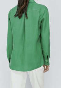 Chemise en lin verte avec col, manches longues et poignets à boutons. Texture lisse, coupe décontractée et ourlet droit. Pas de motifs visibles.