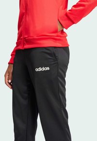 Červená športová mikina na zips s rebrovanými manžetami v kombinácii s čiernymi nohavicami, na ktorých je vyšitý biely logo "adidas". Hladká textúra látky.