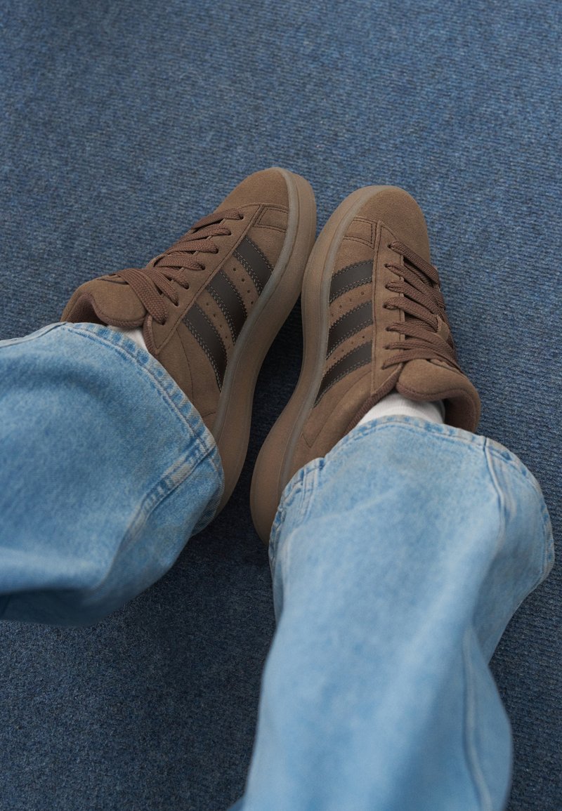 Brown suede sneakers with dark brown stripes, rubber soles, and textured laces, paired with light blue jeans on a blue carpet.