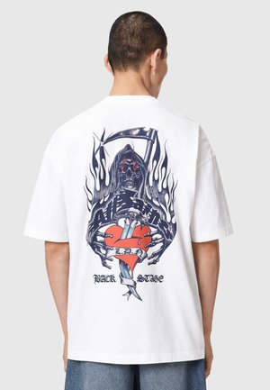Personne portant un t-shirt blanc avec un graphisme d’un squelette à capuche tenant un cœur transpercé par un poignard, des flammes et le texte « L.D.A » et « Back Stage ».