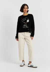 Un pull en maille noir avec une illustration de Snoopy entouré de cœurs blancs et rouges, associé à un pantalon beige et des baskets blanches avec des accents noirs.