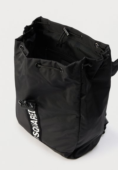 Μαύρο σακίδιο πλάτης από νάιλον με κορδόνι σύσφιξης, μπροστινό λουρί με αγκράφα και λευκό κείμενο "DSQUARED" σε μαύρο κάθετο πλαίσιο.