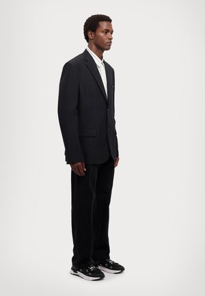 Blazer nero con un bottone singolo, abbinato a una camicia bianca a righe e pantaloni neri a gamba dritta. Sneakers nere completano l'outfit.