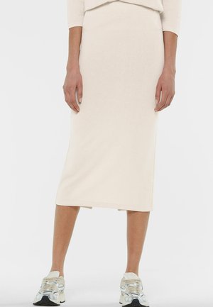 Frau trägt einen cremefarbenen Midirock kombiniert mit weißen und silbernen klobigen Sneakers, steht vor einem schlichten Hintergrund.