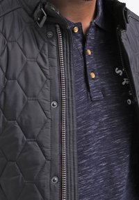 Quiltad svart jacka med dragkedja fram och tryckknappar, lager på en marinblå polo med strukturerad yta och knappar.