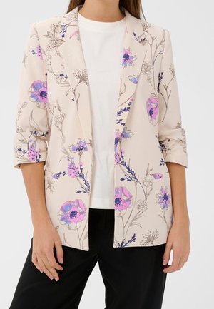 Mujer con blazer beige de mangas remangadas y estampado floral rosa-púrpura sobre blusa blanca y pantalones negros, con las manos relajadas a los lados.