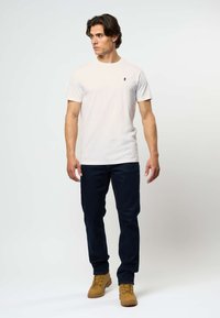 Valkoinen lyhythihainen t-paita, jossa on pieni tummansininen logo, yhdistettynä tummansinisiin farkkuihin ja vaaleanruskeisiin saappaisiin, seisoo yksinkertaista taustaa vasten.