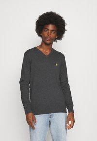 Suéter gris de cuello en V, hecho de material de punto, con mangas largas y un pequeño logo amarillo en el pecho izquierdo. Diseño minimalista y textura suave.