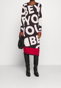 Strickkleid mit langen Ärmeln in Schwarz, Weiß und Rot mit großem "OBEY YOU" Schriftzug. Kombiniert mit kniehohen schwarzen Stiefeln und einer strukturierten schwarzen Tasche.
