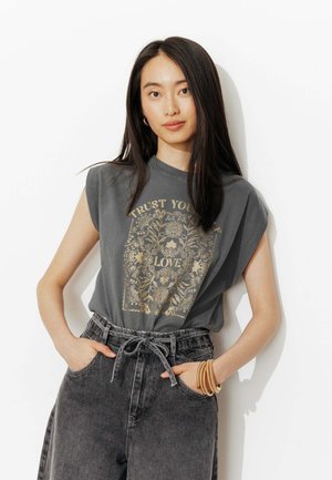 Femme aux longs cheveux noirs portant un T-shirt graphique gris sans manches avec le texte "Fais confiance à ton cœur, aime" et un jean gris foncé taille haute, ainsi que des bracelets.