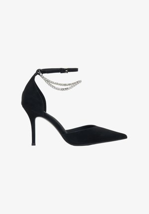 Zwarte suède hoge hak met een spitse neus, enkelbandje en twee kettingen met kristalaccenten. De stilettohak is ongeveer vier inch hoog.