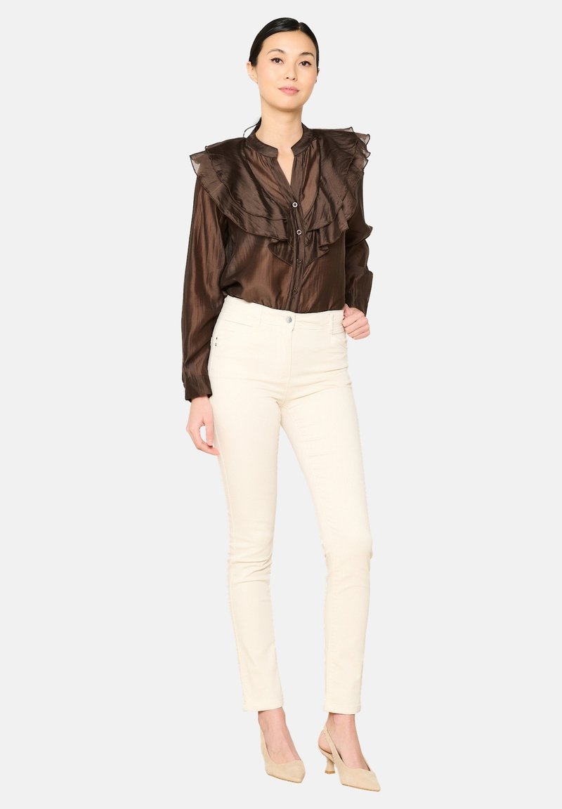 Ruda satino bluzelė su išsiskiriančiu raukšlėtu apykakle, sagčių užsegimas; derinama su šviesiai kreminėmis prigludusiomis kelnėmis ir kūno spalvos aukštakulniais. Neutralus fonas.