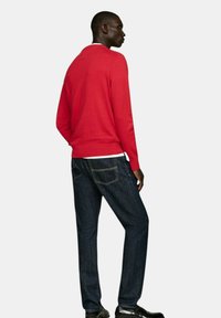 Maglione rosso a maglia con maniche lunghe, abbinato a jeans di denim scuro con cuciture in vista e una vestibilità classica. Le scarpe nere completano l'outfit.