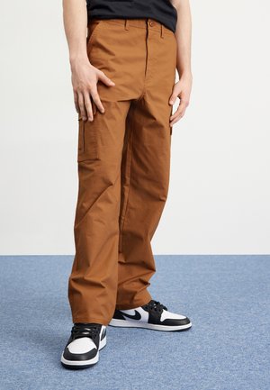Personne portant un pantalon cargo marron et des baskets noires et blanches, debout sur un tapis bleu, les mains le long du corps.