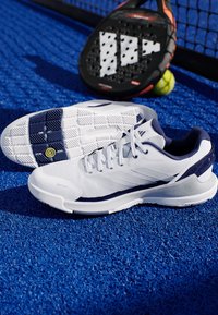 Bílé sportovní boty s námořnickými akcenty, síťovaný svršek, texturovaná podešev a polstrovaná pata, umístěné na modré hrací ploše vedle padelové rakety.