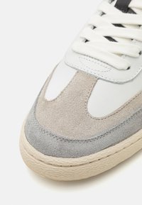 Adidași cu un superior texturat din suede gri și bej, accente din piele albă, sireturi rotunde și o talpă din cauciuc de culoare deschisă.