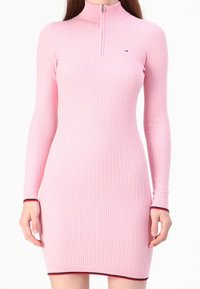 Rosa ribbad klänning med långa ärmar och hög krage. Har en dragkedja fram och kontrasterande röd och vit kant vid ärmslut och fåll.