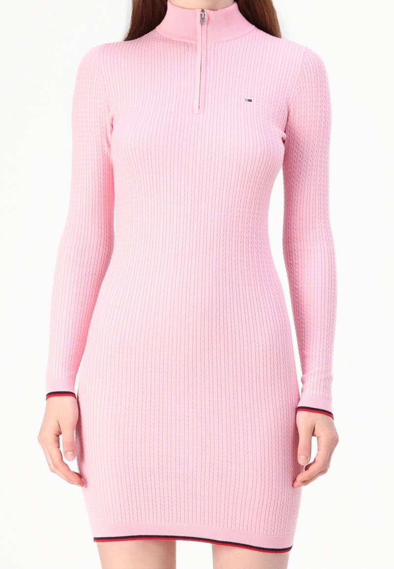 Rosa ribbad klänning med långa ärmar och hög krage. Har en dragkedja fram och kontrasterande röd och vit kant vid ärmslut och fåll.