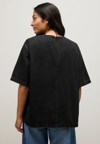 T-shirt en denim noir à manches courtes, coupe décontractée, et ouverture au niveau de la nuque à l'arrière. Le tissu a une texture douce et des coutures minimales.