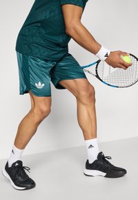 Türkisfarbenes Sportshirt mit strukturiertem Muster, passende türkisfarbene Shorts, weißes Logo, schwarze Schuhe, weiße Knöchelsocken, hält einen Tennisball und einen Schläger.