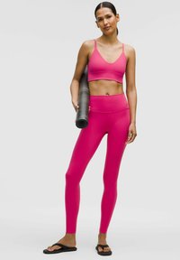Un sutien sport roz strălucitor și colanți asortați, dintr-un material neted și elastic; modelul ține o saltea de yoga neagră, purtând flip-flops negri.