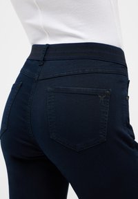 Tmavě modré džíny s hladkou texturou, vybavené širokým elastickým pasem a dvěma zadními kapsami s jemnými švy.