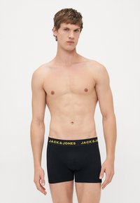 Junger Mann steht in schwarzen Jack & Jones Boxer-Shorts mit gelbem Logo am Bund, mit neutralem Gesichtsausdruck, vor einem schlichten hellen Hintergrund.