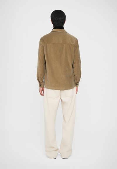 Camicia in corduroy marrone con colletto e yoke posteriore, caratterizzata da maniche lunghe, polsini con bottone e orlo leggermente arrotondato. Abbinata a pantaloni color crema.