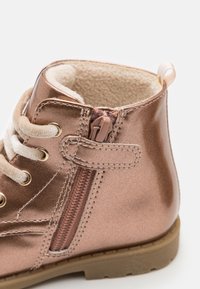 Acercamiento a una bota infantil metálica color bronce con cordones crema, cremallera lateral, forro de felpa suave y suela de goma beige.