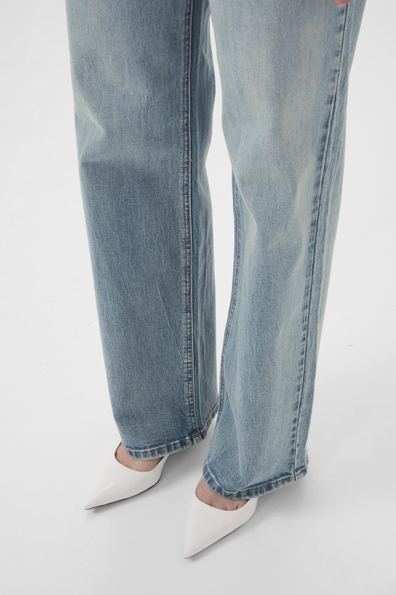 Jean en denim bleu clair à jambes larges, associé à des chaussures blanches à talons hauts et pointues. Le jean présente des coutures apparentes et une coupe décontractée.