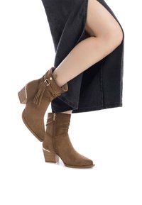 Botas de tobillo de ante marrón con diseño arrugado, cremallera lateral, acento de borlas y un tacón apilado que presenta un detalle metálico en la parte posterior.
