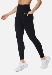 Leggings neri a vita alta con tasca laterale, caratterizzati da cuciture a contrasto e un logo sulla parte anteriore. Abbinati a scarpe da ginnastica bianche.