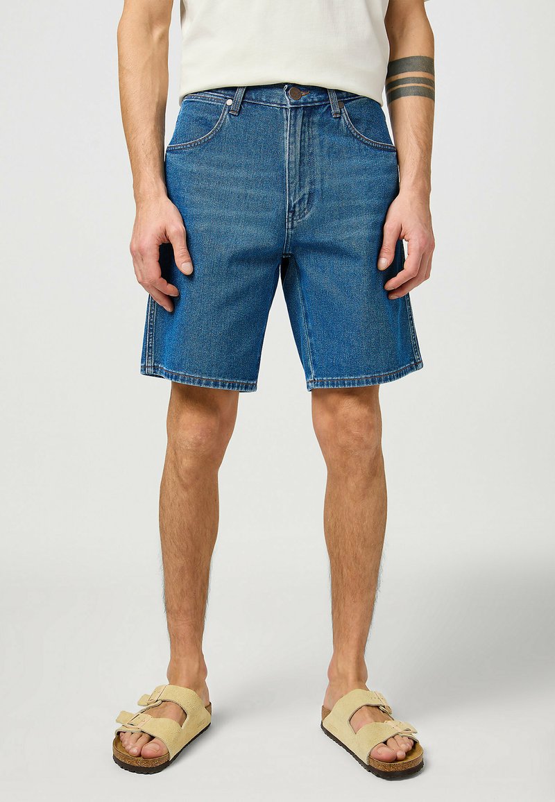 Spijker shorts in medium blauw met een klassieke pasvorm, voorzien van vijfzakken, riemlussen en een onafgebroken zoom. Draagt met beige sandalen met open tenen.